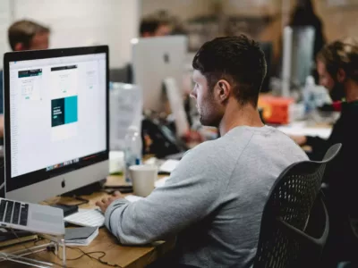 Student sitzt am Tisch vor einem iMac und schaut auf den Bildschirm