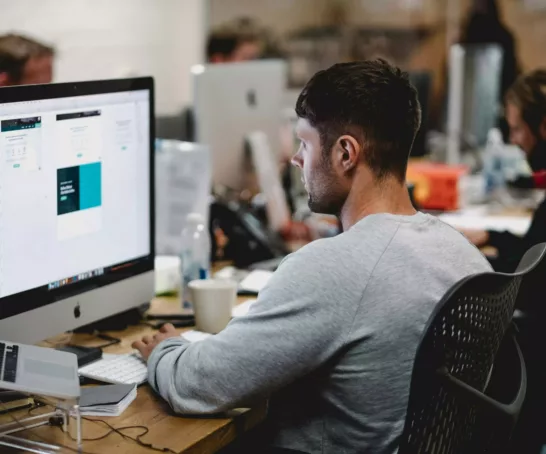 Student sitzt am Tisch vor einem iMac und schaut auf den Bildschirm